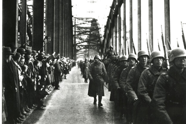 Soldaten der Wehrmacht in Uniform mit Helm marschieren über die Rheinbrücke. Dabei werden sie von vielen Zuschauern auf der anderen Seite der Brücke beobachtet