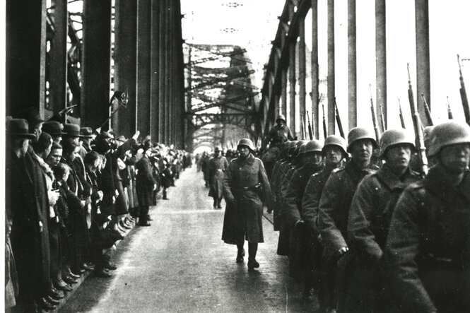 Soldaten der Wehrmacht in Uniform mit Helm marschieren über die Rheinbrücke. Dabei werden sie von vielen Zuschauern auf der anderen Seite der Brücke beobachtet
