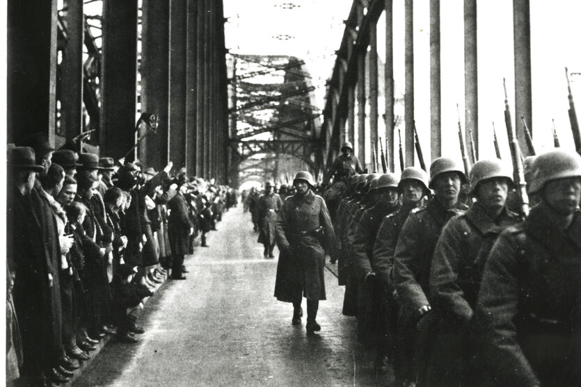 Soldaten der Wehrmacht in Uniform mit Helm marschieren über die Rheinbrücke. Dabei werden sie von vielen Zuschauern auf der anderen Seite der Brücke beobachtet