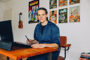 Zu sehen ist die Comic-Zeichnerin Celine Keller an ihrem Schreibtisch. Im Hintergrund eine Wand, die mit bunten Bildern und einer Ukulele behängt ist.