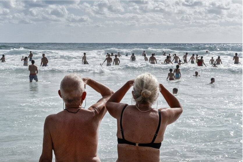 Zwei ältere Badende blicken auf ein Meer mit vielen Touristen im Wasser.
