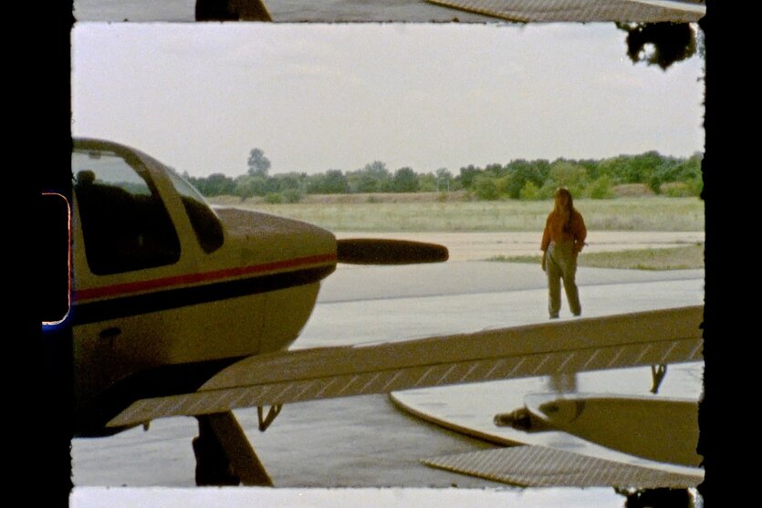 Ein Filmbild mit einer Frau vor einem Propellerflugzeug.