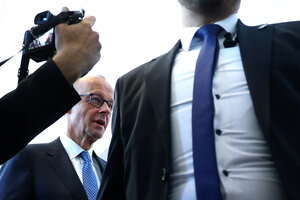 Ein Mann mit breiter Brust in Hemd und Krawatte steht vor Friedrich Merz, der hinter ihm geht. Ein Fotograf richtet seine Kamera auf ihn, zu sehen ist nur die Kamera und der Unterarm