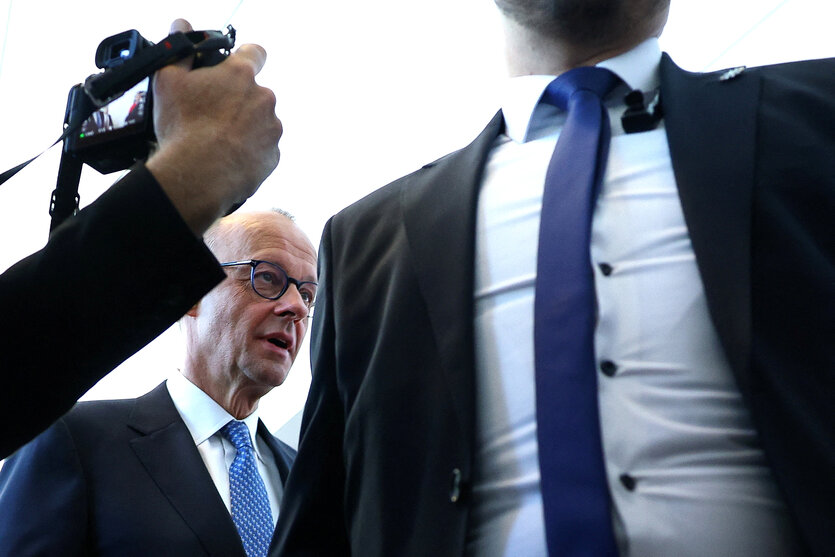 Ein Mann mit breiter Brust in Hemd und Krawatte steht vor Friedrich Merz, der hinter ihm geht. Ein Fotograf richtet seine Kamera auf ihn, zu sehen ist nur die Kamera und der Unterarm