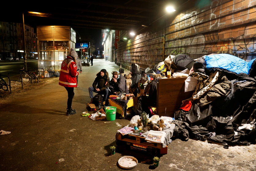 Eine Person in der roten Jacke des Roten Kreuzes steht vor Menschen, die auf einem Gehweg sitzen, es liegt Schnee