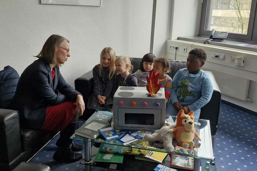 Bildungssenatorin Sascha Aulepp sitzt mit vier Kitakindern an einem Tisch; in der MItte ein gebastelter Pappherd, der in Flammen aufgeht