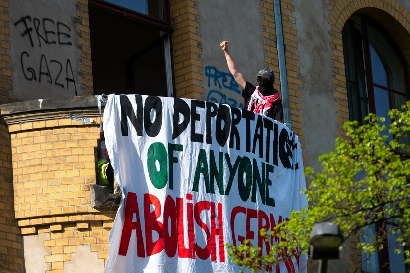 Ein Aktivist hebt die Faust auf einem Balkon in einem Gebäude am Campus Nord der Humboldt-Universität (HU) und steht neben einem Banner mit Forderungen gegen Abschiebungen