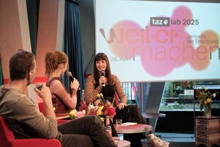 Ein Bild einer Veranstaltung des taz labs 2025. Es sind drei Personen auf einem Podium zu sehen, Susanne Schwarz, Carla Hinrichs und Tadzio Müller. im Hintergrund steht auf einem Bildschirm "taz lab 2025. Weitermachen. Jenseits der Empörung. 26. April."