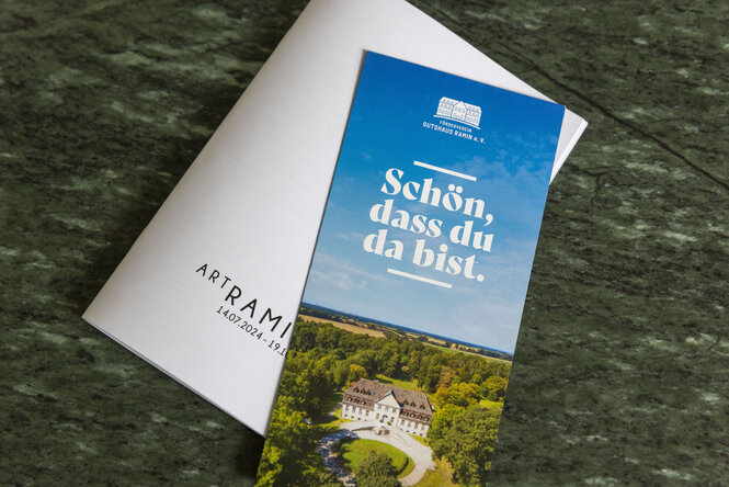 Ein Faltblatt mit dem Text „Schön, dass du da bist“ und einem Foto eines Gutshauses im Grünen liegt auf einer grünen Marmoroberfläche, daneben ein weißes Kuvert mit der Aufschrift „ARTAMI“ und einem Datum.