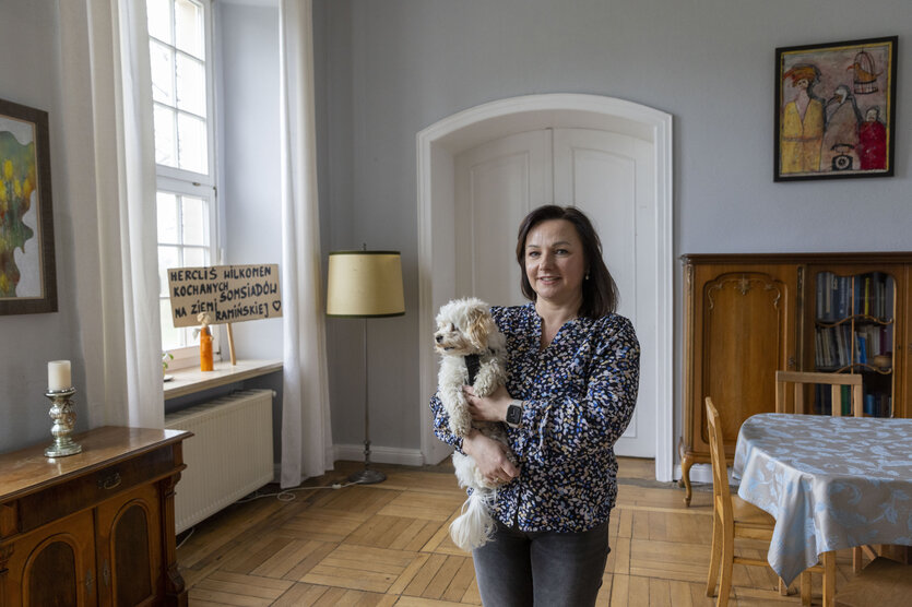 Eine Frau mit einem kleinen weißen Hund steht in einem hellen Raum mit Holzboden, großen Fenstern und einem polnischen Willkommensschild im Hintergrund