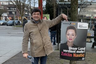 Ein Mann in beiger Jacke zeigt auf ein Plakat