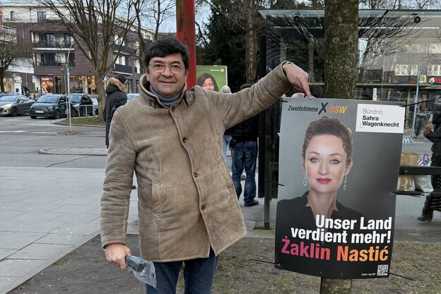 Ein Mann in beiger Jacke zeigt auf ein Plakat