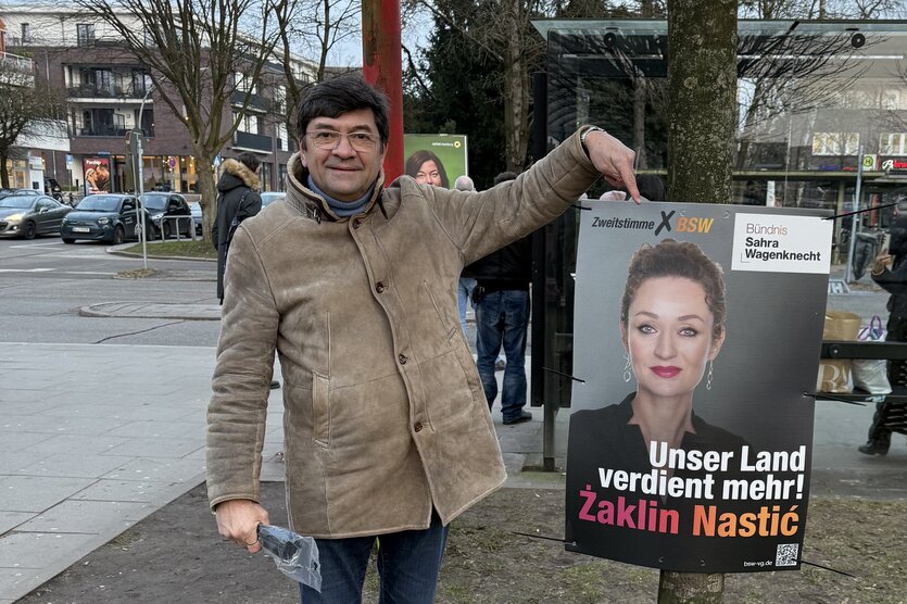 Ein Mann in beiger Jacke zeigt auf ein Plakat