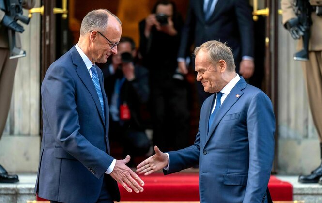 Friedrich Merz und Donald Tusk trefffen sich - die Hände sind ausgestreckt, aber noch berühren sie sich nicht. Beide Männer lächeln verhalten