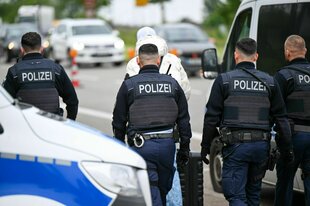 Rückenansicht von vier Polizisten, die Handschellen am Gürtel tragen, gehen vor einem Mann, der ein helle Jacke trägt