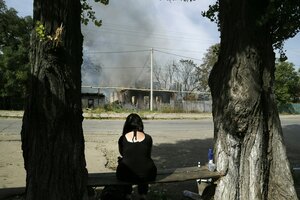 Eine Person sitzt vor einem brennenden Haus und sieht wie die Rauchschwaden aufsteigen