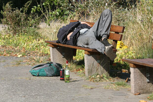 Ein Mann liegt auf einer Parkbank, daneben liegt ein Rucksack und steht eine Flasche Wein
