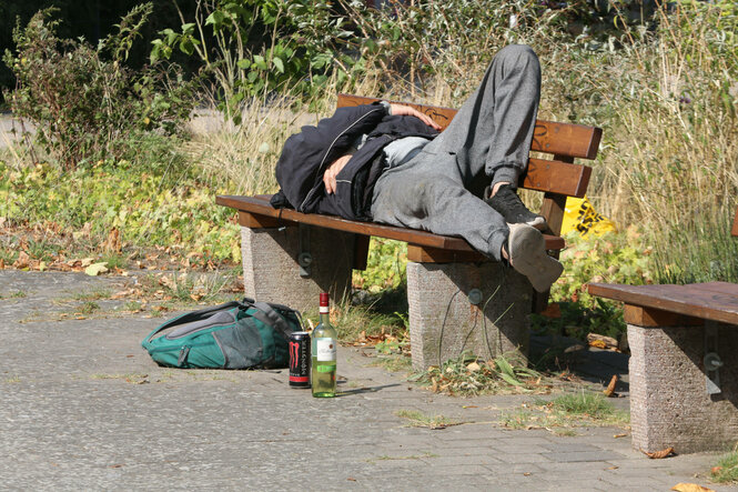 Ein Mann liegt auf einer Parkbank, daneben liegt ein Rucksack und steht eine Flasche Wein