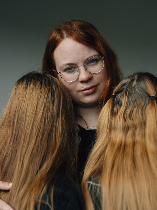 Drei Personen stehen eng zusammen, eine Person schaut in die Kamera, zwei stehen abgewandt vor ihr