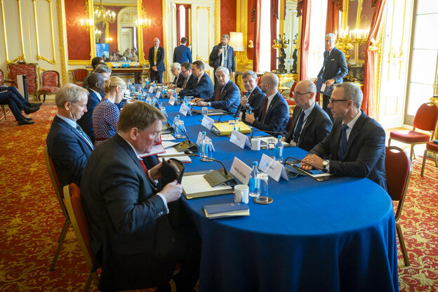 In einem prunkvollem Saal sitzen viele Männer und eine Frau an einem Tisch mit blauer Tischdecke