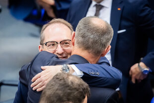 Merz und Dobrindt umarmen sich im Deutschen Bundestag. Sie tragen Anzüge. Dobrindt ist dem Betrachter zugewandt, lächelt zufrieden und trägt eine Brille. Von Merz sieht man den Hinterkopf.