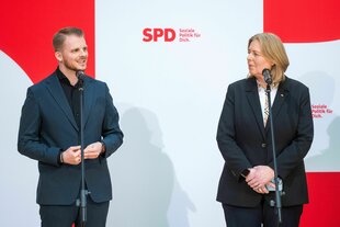 Tim Klüssendorf und Bärbel Bas schauen sich gegenseitig an und stehen vor einer Plakatwand mit dem SPD-Parteilogo