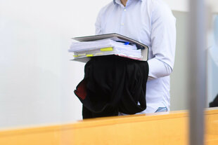 Zeigt den Angeklagten als er zu seinem Platz im Gerichtssaal geht. Zu sehen sind nur sein Oberkörper in einem weißen Hemd und seine vor dem Bauch mit Handschellen gefesselten Hände, über die ein schwarzes Jackett hängt. Darauf balanciert er einen dicken Aktenordner.