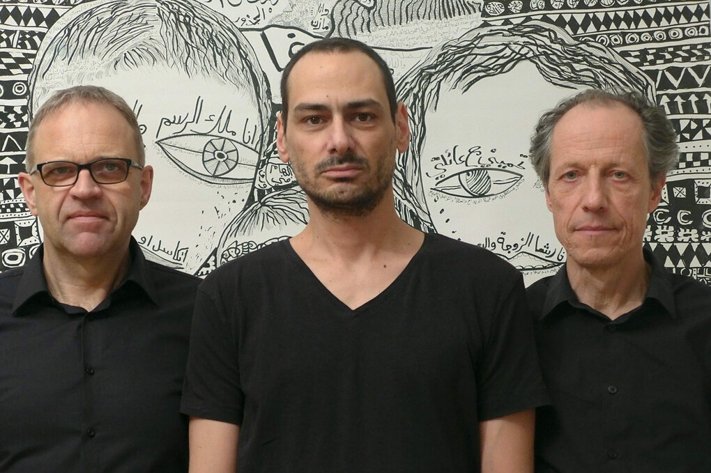 Das Trio Sawt Out steht vor einer Schwarz-Weiß-Zeichnung von Mazen Kerbaj. Die drei Männer tragen schwarze T-Shirts. Die Zeichnung im Hintegrund ist Wandgroß und lässt zwei Gesichter mit großen Augen erkennen.