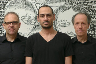 Das Trio Sawt Out steht vor einer Schwarz-Weiß-Zeichnung von Mazen Kerbaj. Die drei Männer tragen schwarze T-Shirts. Die Zeichnung im Hintegrund ist Wandgroß und lässt zwei Gesichter mit großen Augen erkennen.