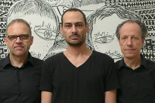Das Trio Sawt Out steht vor einer Schwarz-Weiß-Zeichnung von Mazen Kerbaj. Die drei Männer tragen schwarze T-Shirts. Die Zeichnung im Hintegrund ist Wandgroß und lässt zwei Gesichter mit großen Augen erkennen.