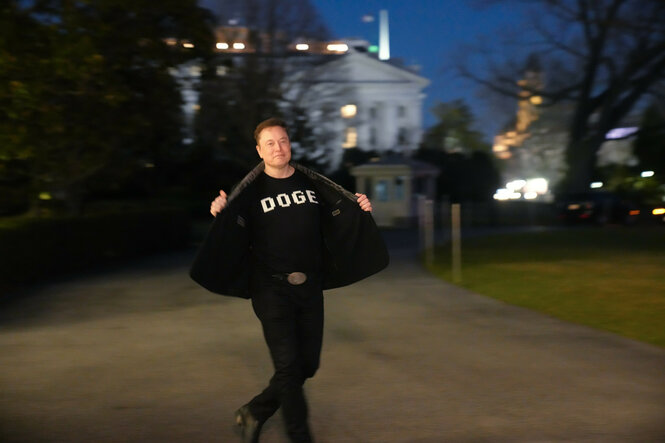 Zu sehen ist Elon Musk. Er zeigt den Betrachter:innen sein T-Shirt mit der Aufschrift "DOGE", während er auf dem Südrasen des Weißen Hauses in Washington geht. DOGE steht für das Department of Government Efficiency.