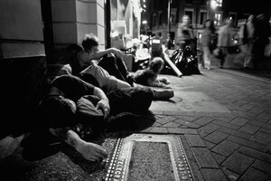 Jugendliche sitzen und schlafen auf einem Bürgersteig, Passanten laufen vorbei