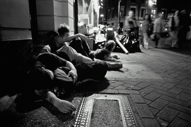 Jugendliche sitzen und schlafen auf einem Bürgersteig, Passanten laufen vorbei