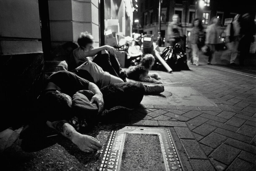 Jugendliche sitzen und schlafen auf einem Bürgersteig, Passanten laufen vorbei