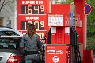 Eine Frau hält an einer Tankstelle vor einer Preistafel einen Tankstutzen.