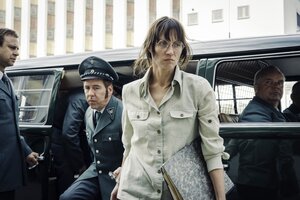 Eine Frau mit Brille und Aktenordner unterm Arm steht vor einem Polizeiwagen, zwei Polizisten hinter ihr