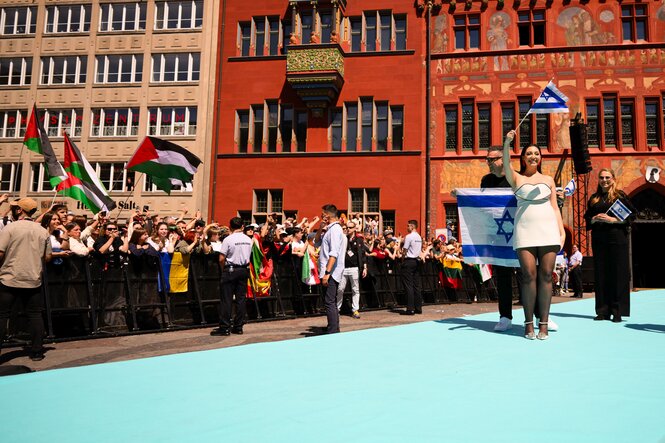 Yuval Raphael tritt vor Altbauten in Basel auf einem Teppich mit israelischer Fahne auf, links von ihr Protestierende mit palästinensischen Fahnen