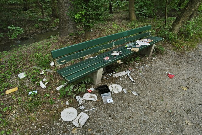 Plastikteller, Pappschalen und anderer Müll liegen auf und neben einer grünen Parkbank.