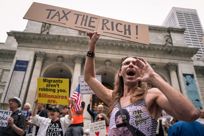 Ein junge, männlich gelesene Person mit langen Haaren und Tanktop hält vor einer demonstrierenden Menge ein Pappschild hoch, auf dem steht "Tax the Rich" – er scheint etwas zu rufen und formt eine Hand vor seinem Mund zu einem Lautsprecher.