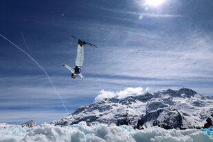 Ein Freestyle-Skiläufer steht kopfüber in der Luft, hinter ihm die schweizer Berge