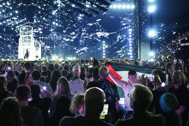 ein publikum vor einer erleuchteten bühne, darunter ein mann mit palästina flagge