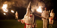 Mitglieder des rassistischen Ku-Klux Klans wandern mit Fackeln und weißen Mützen im Kreis um ein brennendes Kreuz.