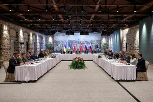 Personen sitzen an einem U-förmigen Tisch, im Hintergrund Fahnen der Ukraine und Russlands, in der Mitte die der Türkei