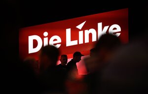 Das rote Logo der Linken leuchtet in der Dunkelheit einer Halle, man sieht die dunklen Silhouetten einiger Menschen