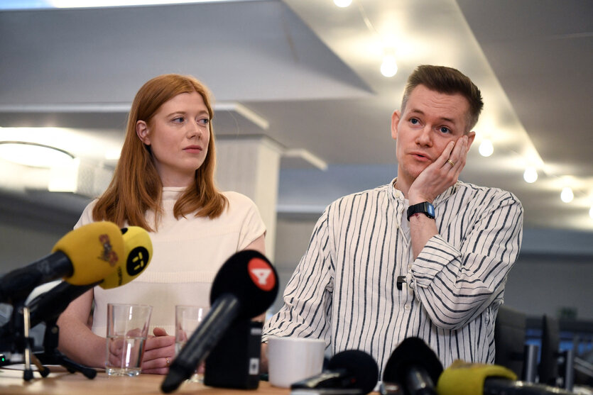 Joakim Medin und seine Frau Sofie Axelsson stehen hinter einem Pult mit Mikrofonen. Joakim hält sich seine rechte Hand an die Wange