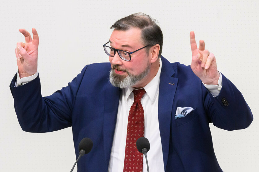 Klaus Wichmann in blauben Jackett und rotem Schlips malt mit den Findern Anführungszeichen in die Luft