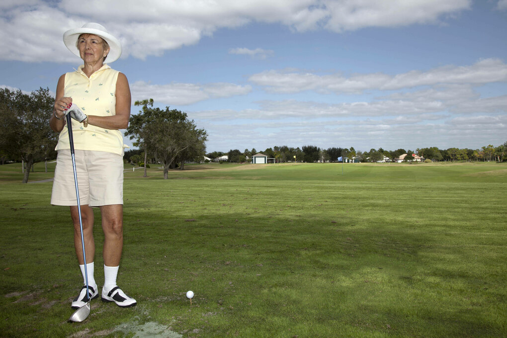 eine ältere Frau mit Golfschläger und in Golfklamotten auf dem grünen Rasen