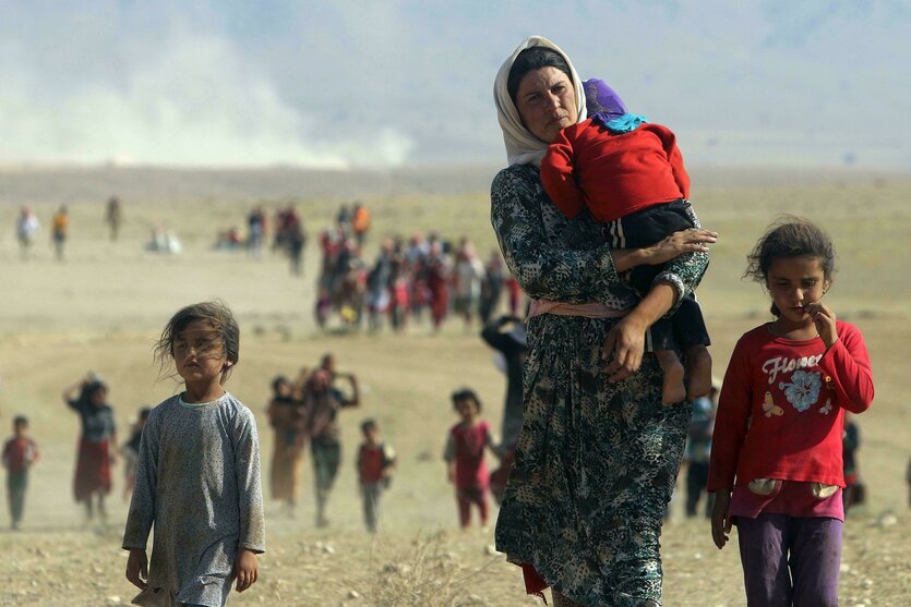 Eine Frau trägt ein Kleinkind auf dem Arm und läuft zusammen mit anderen Kindern durch eine wüstenähnliche Landschaft, weitere Personen folgen in der Ferne