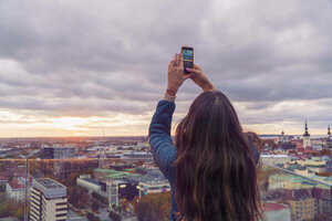 Rückenansicht einer Person, die ihr Smartphone in die Höhe streckt, vor ihr die Skyline von Tallinn