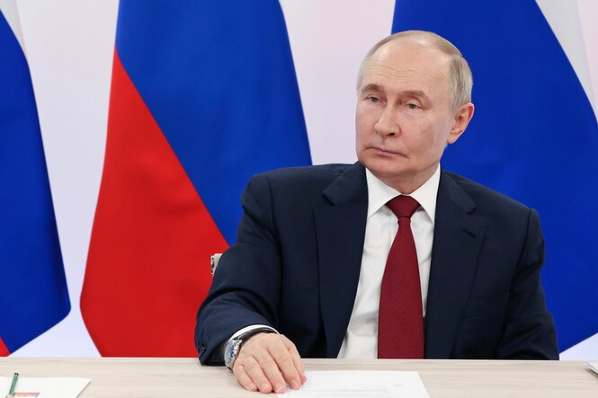 Wladimir Putin sitzt an einem Tisch. Hinter ihm sind die Farben der russischen Flagge zu sehen.